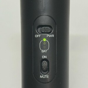May include: Close-up of a black microphone with control buttons. The buttons include "OFF", "PWR", "BAT", "ON", and "MUTE". A green light indicates the power status. The microphone is designed for audio recording or live performances.