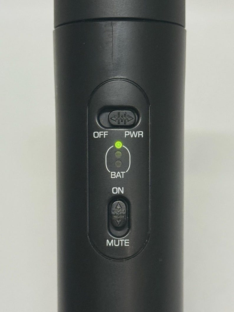 May include: Close-up of a black microphone with control buttons. The buttons include "OFF", "PWR", "BAT", "ON", and "MUTE". A green light indicates the power status. The microphone is designed for audio recording or live performances.