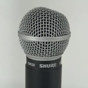 May include: A close-up of a Shure SM58 microphone. The microphone has a silver wire mesh head and a black cylindrical body. The brand name "SHURE" is printed in white on the body.