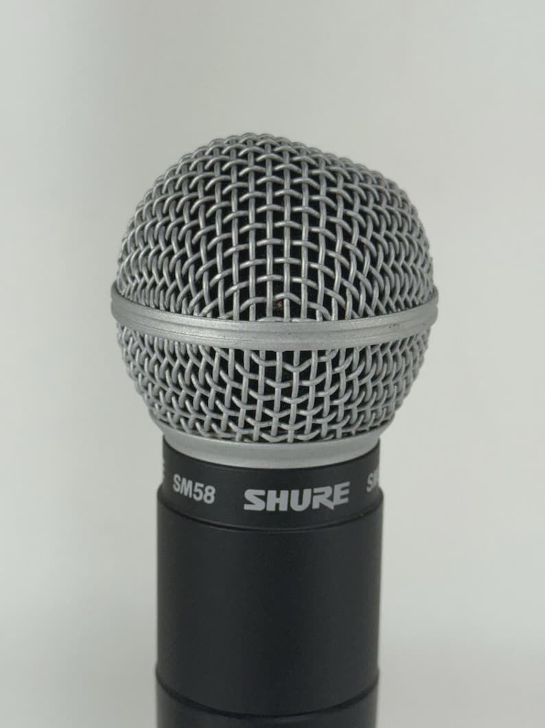 May include: A close-up of a Shure SM58 microphone. The microphone has a silver wire mesh head and a black cylindrical body. The brand name "SHURE" is printed in white on the body.