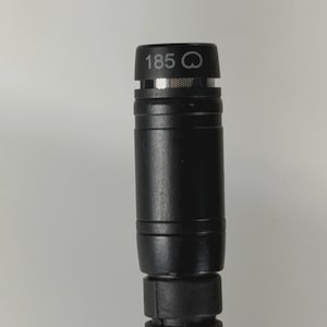 May include: A black cylindrical microphone with a metallic mesh grille at the top. The top section features the printed text 185 followed by a small circular icon. The body has a smooth finish with a ribbed, flexible section at the bottom.