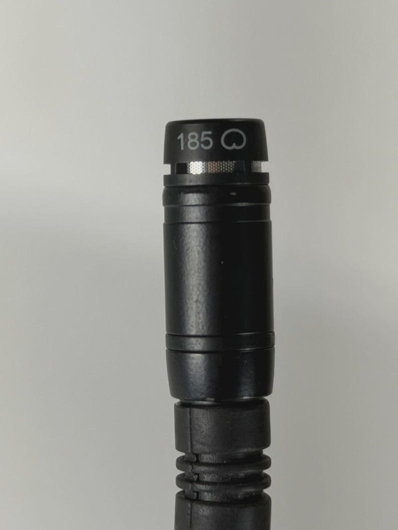 May include: A black cylindrical microphone with a metallic mesh grille at the top. The top section features the printed text 185 followed by a small circular icon. The body has a smooth finish with a ribbed, flexible section at the bottom.