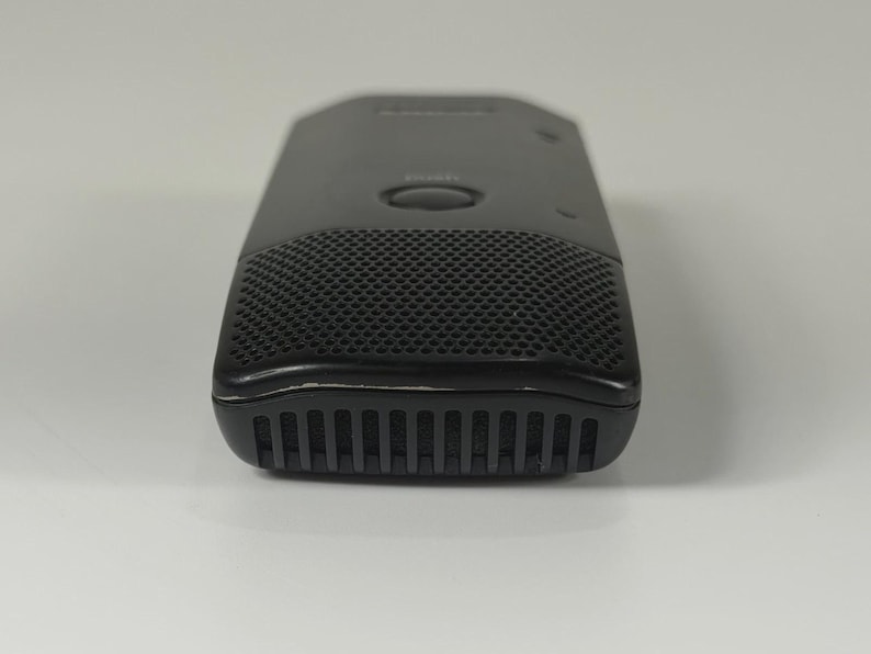 May include: A black rectangular digital voice recorder featuring a perforated speaker grille on the front, a central circular button, and a series of vertical vents along the bottom edge. The device has a smooth, matte finish with subtle branding.