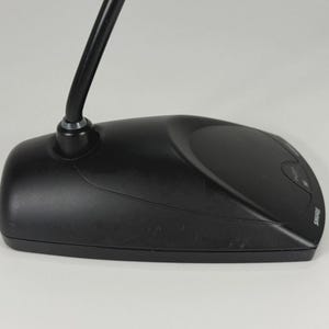 May include: A black desktop microphone base with a curved neck extending upwards. The base features a smooth, matte finish, a prominent push button for operation, and the brand name SHURE printed in white lettering on the right side of the unit.
