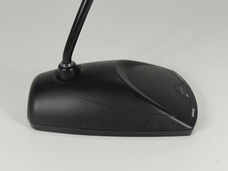 May include: A black desktop microphone base with a curved neck extending upwards. The base features a smooth, matte finish, a prominent push button for operation, and the brand name SHURE printed in white lettering on the right side of the unit.