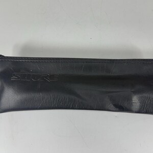 May include: Black leather pouch with the embossed brand name "SHURE". The pouch is rectangular with a zipper closure. The leather has a textured appearance.