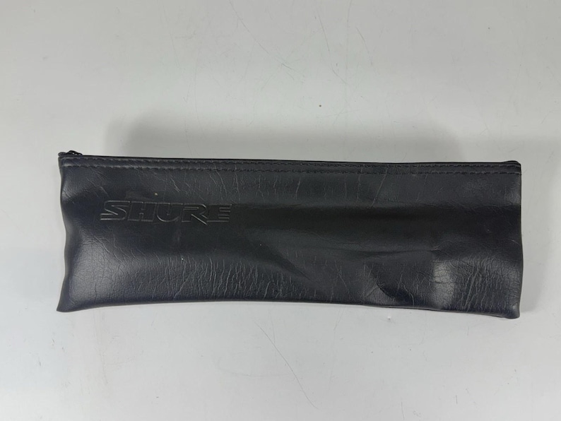 May include: Black leather pouch with the embossed brand name "SHURE". The pouch is rectangular with a zipper closure. The leather has a textured appearance.