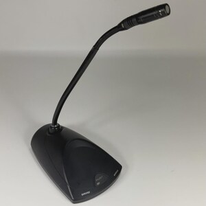 May include: A black Shure gooseneck microphone featuring a flexible neck and a sturdy base. The base includes a push button for operation and the brand name Shure printed in white. The microphone head is cylindrical and positioned at an angle.