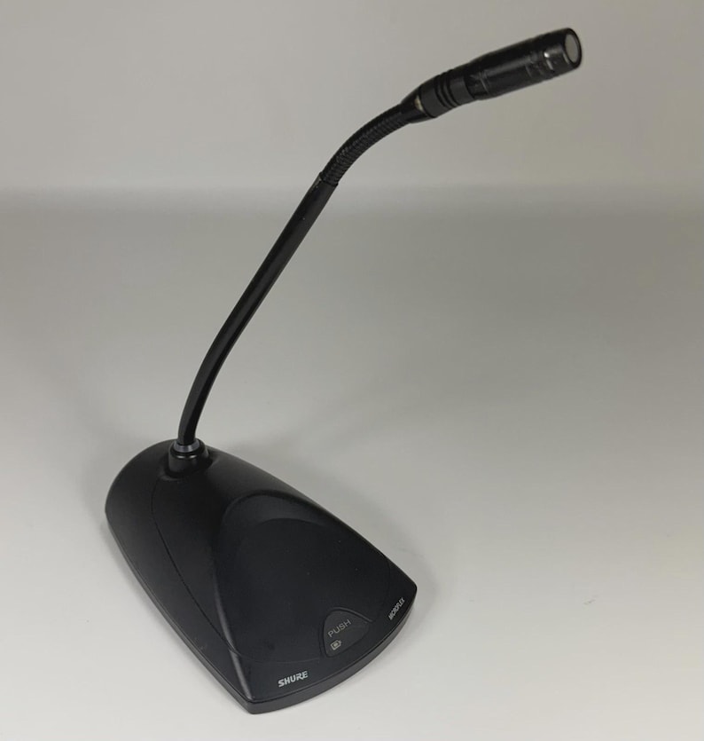 May include: A black Shure gooseneck microphone featuring a flexible neck and a sturdy base. The base includes a push button for operation and the brand name Shure printed in white. The microphone head is cylindrical and positioned at an angle.