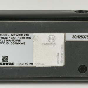 May include: A black Shure MXW6/C Z10 microphone transmitter featuring a white label with technical specifications including frequency 1920-1930 MHz, IC: 616A-MXW6, FCC ID: DD4MXW6, input 5V, a QR code, and a serial number 3QH2537659 on a separate label.