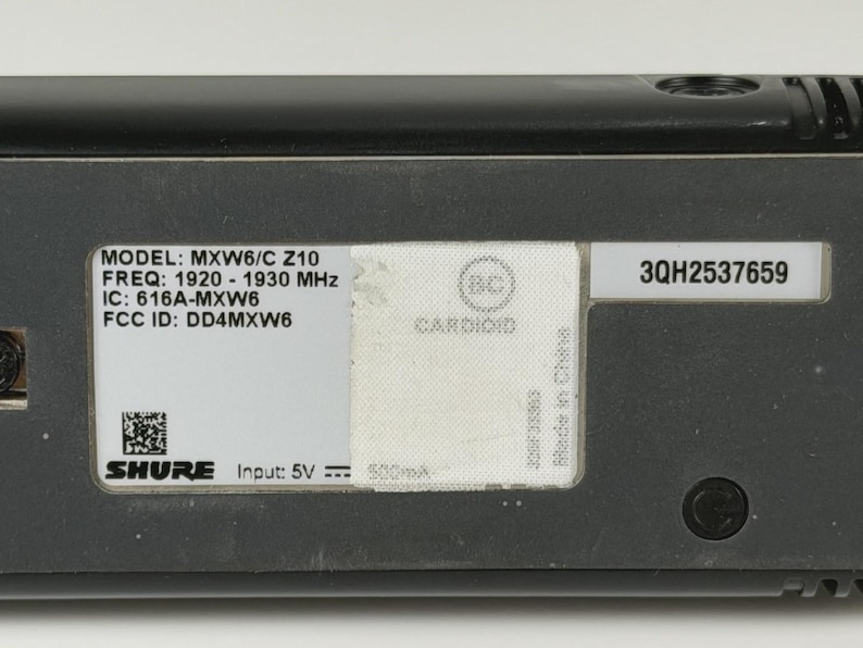 May include: A black Shure MXW6/C Z10 microphone transmitter featuring a white label with technical specifications including frequency 1920-1930 MHz, IC: 616A-MXW6, FCC ID: DD4MXW6, input 5V, a QR code, and a serial number 3QH2537659 on a separate label.