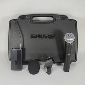 Puede incluir: Un sistema de micr&oacute;fono inal&aacute;mbrico Shure negro con un estuche de transporte. El juego incluye un micr&oacute;fono de mano, un receptor, un clip para micr&oacute;fono y un parabrisas. El logotipo de Shure es visible en el estuche y el receptor.