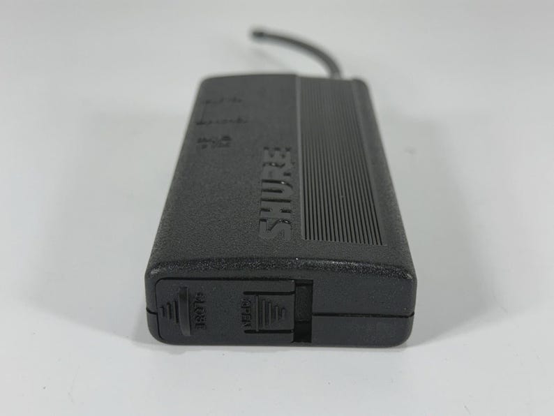 Puede incluir: Un receptor de micr&oacute;fono inal&aacute;mbrico Shure negro con una parte superior acanalada y el nombre de la marca impreso en el lateral. El receptor tiene un peque&ntilde;o cable negro adjunto y un interruptor con la etiqueta "OPEN".