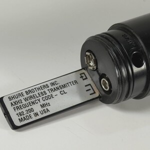 以下が含まれることがあります： 銀色のバッテリートレイが引き出された黒いShure Brothers Inc. AXH2ワイヤレストランスミッター。ラベルには「SHURE BROTHERS INC. AXH2 WIRELESS TRANSMITTER FREQUENCY CODE - CL 192.200 MHz MADE IN USA」と記載されています。デバイスにはゲインコントロールポートが付いています。