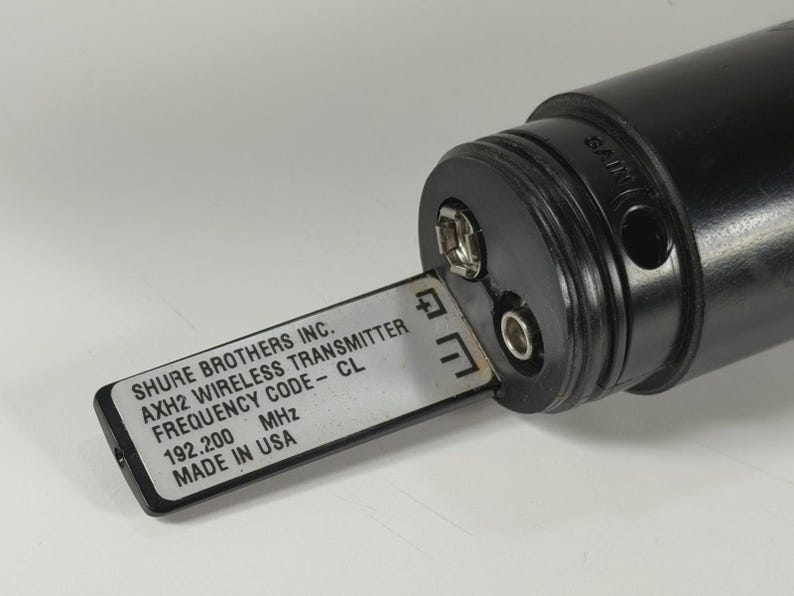 以下が含まれることがあります： 銀色のバッテリートレイが引き出された黒いShure Brothers Inc. AXH2ワイヤレストランスミッター。ラベルには「SHURE BROTHERS INC. AXH2 WIRELESS TRANSMITTER FREQUENCY CODE - CL 192.200 MHz MADE IN USA」と記載されています。デバイスにはゲインコントロールポートが付いています。