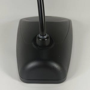 May include: A black desktop microphone base with a smooth, matte finish and a curved, flexible neck extending upwards from the centre. The base has a tapered, rectangular shape with rounded edges and a small grey ring at the base of the neck.