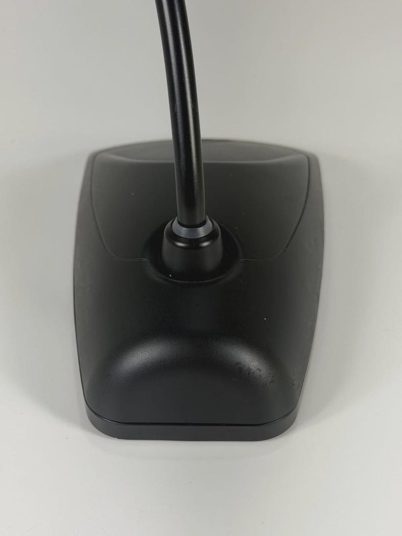 May include: A black desktop microphone base with a smooth, matte finish and a curved, flexible neck extending upwards from the centre. The base has a tapered, rectangular shape with rounded edges and a small grey ring at the base of the neck.