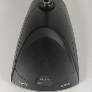 May include: A black Shure Microflex desktop microphone base with a triangular shape. The device features a central button labelled PUSH with a small square icon above it. The brand name SHURE is printed on the left and MICROFLEX on the right.