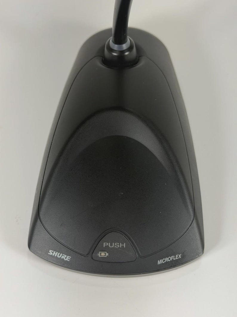 May include: A black Shure Microflex desktop microphone base with a triangular shape. The device features a central button labelled PUSH with a small square icon above it. The brand name SHURE is printed on the left and MICROFLEX on the right.