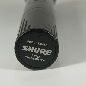 以下が含まれることがあります： 円筒形で垂直な溝がある黒いShure AXH2送信機マイクベース。底部のキャップには、白い文字で「FCC ID: DD4T2」、「SHURE」、「AXH2 TRANSMITTER」と印刷されており、円形の黒い表面の中央に配置されています。