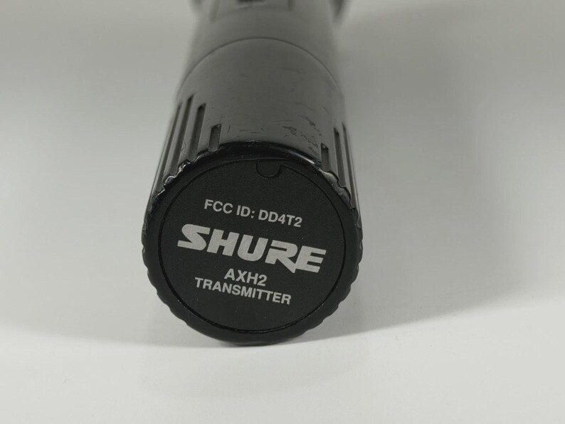 以下が含まれることがあります： 円筒形で垂直な溝がある黒いShure AXH2送信機マイクベース。底部のキャップには、白い文字で「FCC ID: DD4T2」、「SHURE」、「AXH2 TRANSMITTER」と印刷されており、円形の黒い表面の中央に配置されています。