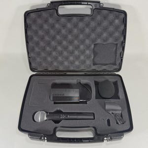 Puede incluir: Maleta de transporte negra abierta que contiene un sistema de micr&oacute;fono inal&aacute;mbrico. La maleta incluye un micr&oacute;fono, un receptor, un parabrisas y un clip para micr&oacute;fono, todo ello encajado en espuma cortada a medida. El micr&oacute;fono es plateado y negro.