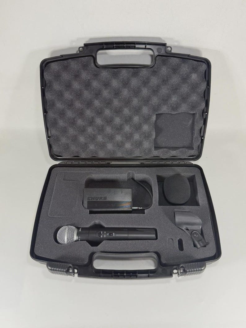 Puede incluir: Maleta de transporte negra abierta que contiene un sistema de micr&oacute;fono inal&aacute;mbrico. La maleta incluye un micr&oacute;fono, un receptor, un parabrisas y un clip para micr&oacute;fono, todo ello encajado en espuma cortada a medida. El micr&oacute;fono es plateado y negro.