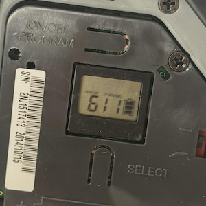 May include: A dark grey electronic device control panel featuring an LCD screen displaying the text GROUP 6, CHANNEL 1, and a battery icon. The panel includes the text ON/OFF PROGRAM, SELECT, and SYNC, along with a white serial number label 2NJ1517413.
