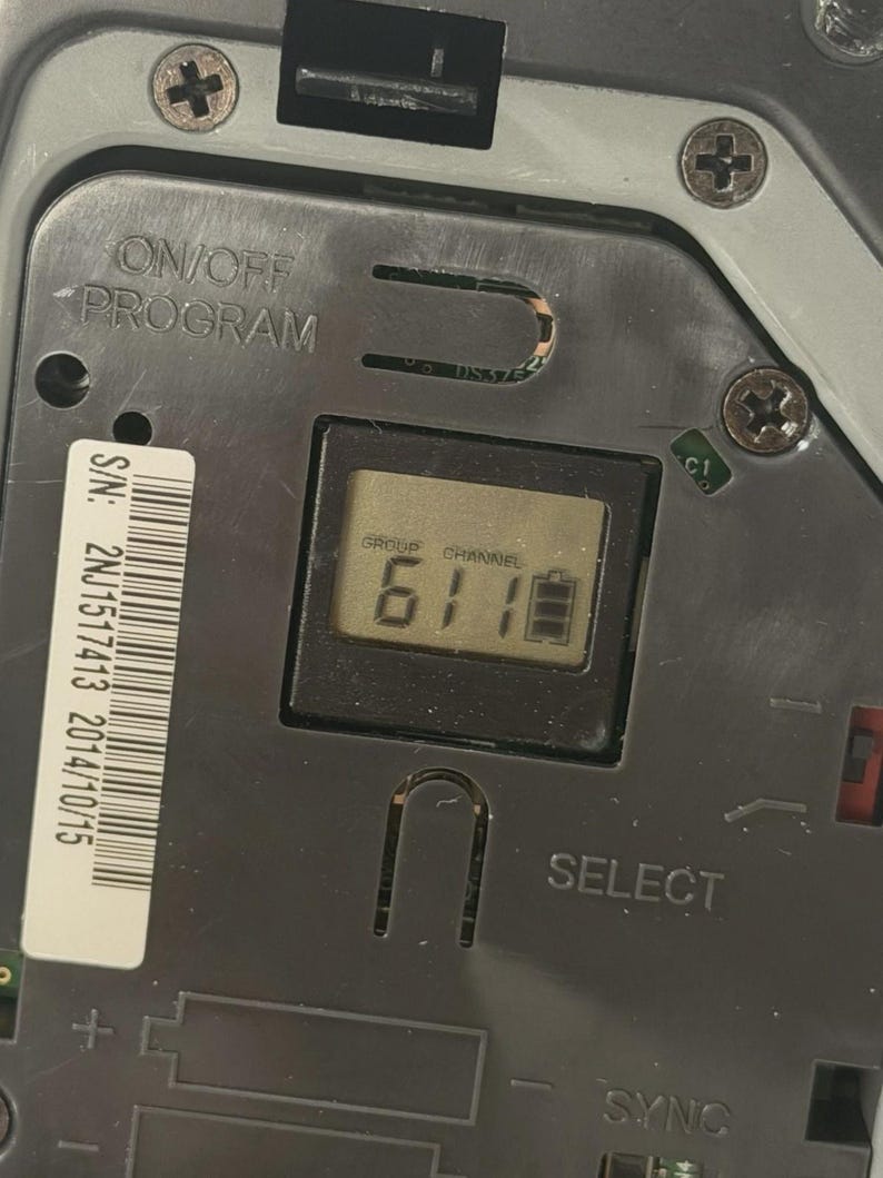 May include: A dark grey electronic device control panel featuring an LCD screen displaying the text GROUP 6, CHANNEL 1, and a battery icon. The panel includes the text ON/OFF PROGRAM, SELECT, and SYNC, along with a white serial number label 2NJ1517413.