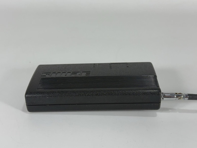 Puede incluir: Un dispositivo de audio Shure negro con una parte superior acanalada y un conector plateado. El dispositivo es rectangular y tiene el nombre de la marca impreso en la parte superior. Un cable negro se extiende desde el conector.