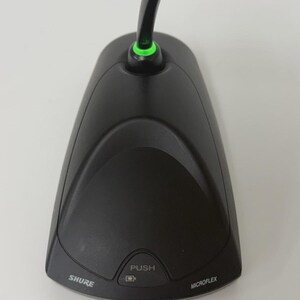 May include: A black Shure Microflex desktop boundary microphone featuring a curved base, a central push button with a small icon, and a green LED light ring at the base of the microphone neck. The brand name Shure and the model name Microflex are printed on the front.