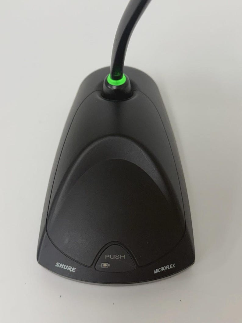 May include: A black Shure Microflex desktop boundary microphone featuring a curved base, a central push button with a small icon, and a green LED light ring at the base of the microphone neck. The brand name Shure and the model name Microflex are printed on the front.