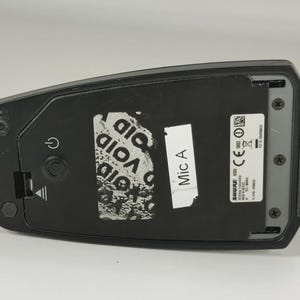 May include: A black Shure wireless transmitter featuring a power button, a battery compartment cover, and a label marked Mic A. The device includes a silver sticker with technical specifications, regulatory symbols, and the text Made in Mexico.