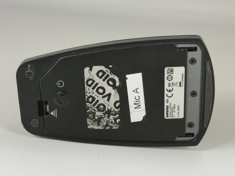 May include: A black Shure wireless transmitter featuring a power button, a battery compartment cover, and a label marked Mic A. The device includes a silver sticker with technical specifications, regulatory symbols, and the text Made in Mexico.
