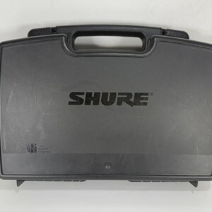 Puede incluir: Una maleta de transporte rectangular negra con asa y el nombre de la marca "SHURE" impreso en letras grandes y en negrita. La maleta est&aacute; dise&ntilde;ada para proteger equipos electr&oacute;nicos.