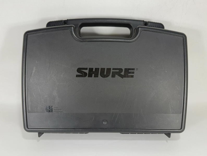 Puede incluir: Una maleta de transporte rectangular negra con asa y el nombre de la marca "SHURE" impreso en letras grandes y en negrita. La maleta est&aacute; dise&ntilde;ada para proteger equipos electr&oacute;nicos.