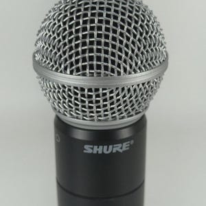 Puede incluir: Primer plano de un micr&oacute;fono Shure. El micr&oacute;fono tiene una cabeza de malla de alambre plateada y un cuerpo negro. La palabra "SHURE" est&aacute; impresa en blanco en el cuerpo del micr&oacute;fono.