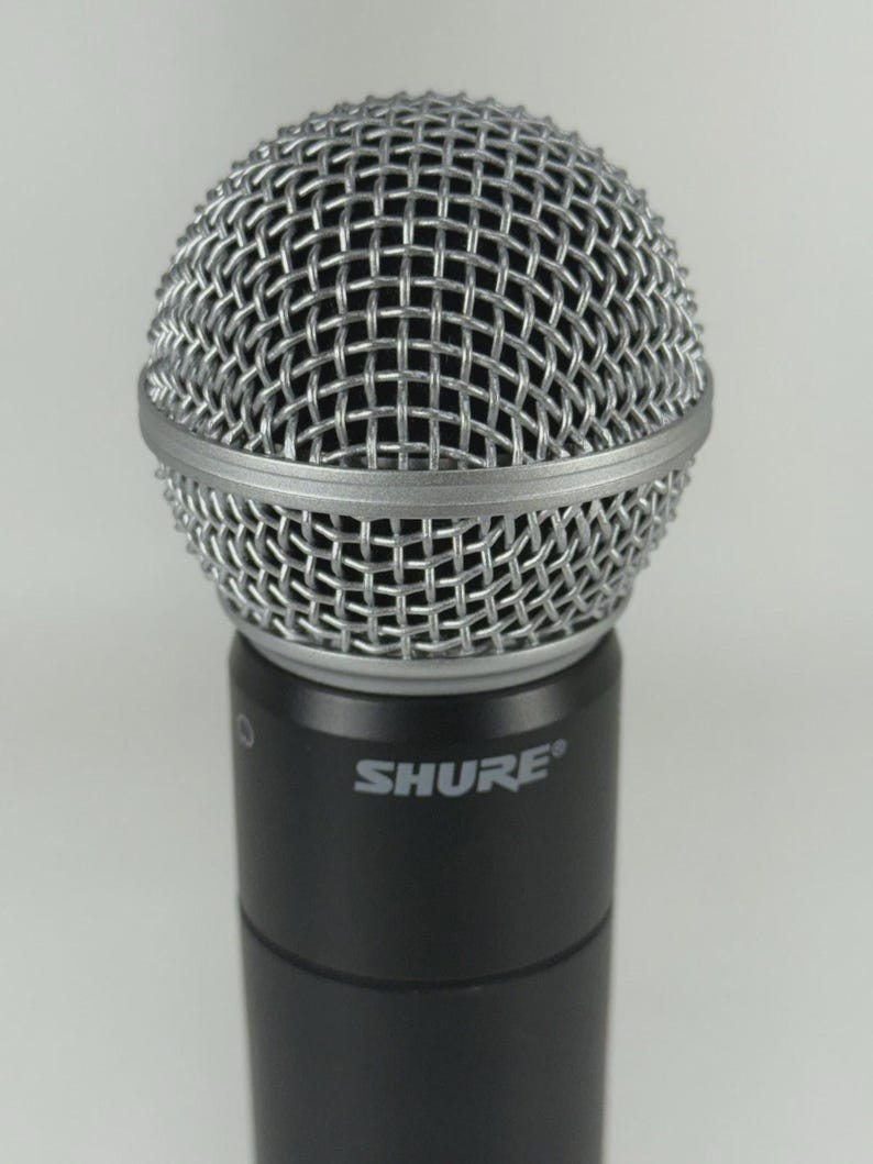 Puede incluir: Primer plano de un micr&oacute;fono Shure. El micr&oacute;fono tiene una cabeza de malla de alambre plateada y un cuerpo negro. La palabra "SHURE" est&aacute; impresa en blanco en el cuerpo del micr&oacute;fono.