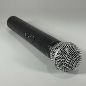 Puede incluir: Un micr&oacute;fono de mano negro con una cabeza de malla plateada. El micr&oacute;fono tiene un cuerpo cil&iacute;ndrico con botones de control y el n&uacute;mero de modelo SM58. El micr&oacute;fono est&aacute; ligeramente inclinado hacia la derecha.