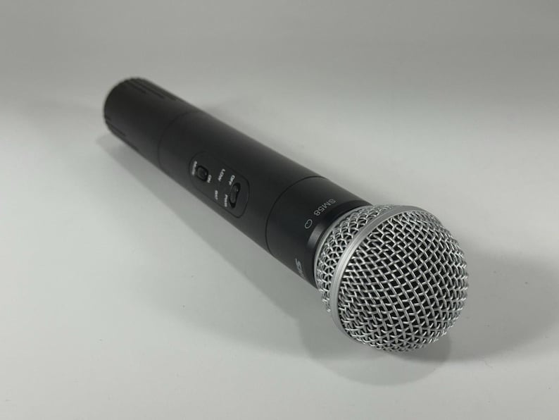 Puede incluir: Un micr&oacute;fono de mano negro con una cabeza de malla plateada. El micr&oacute;fono tiene un cuerpo cil&iacute;ndrico con botones de control y el n&uacute;mero de modelo SM58. El micr&oacute;fono est&aacute; ligeramente inclinado hacia la derecha.