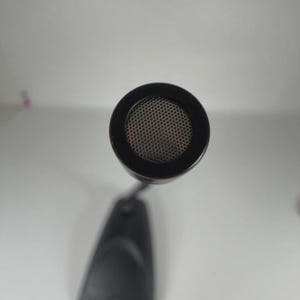 May include: A black gooseneck microphone featuring a circular head with a fine metal mesh grille. The microphone is attached to a solid, tapered black base that sits flat. The view is from directly above, highlighting the circular mesh pattern.