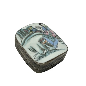 May include: A rectangular, antique trinket box with a silver-colored metal frame. The lid features a hand-painted scene with figures, flowers, and an elephant in shades of blue, pink, and green. The box is likely made of porcelain.