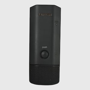 May include: A black Shure wireless microphone transmitter featuring a rectangular body with a central circular push button, a small indicator light, and a perforated metal grille at the bottom. The word push is printed above the button.