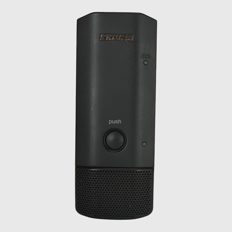 May include: A black Shure wireless microphone transmitter featuring a rectangular body with a central circular push button, a small indicator light, and a perforated metal grille at the bottom. The word push is printed above the button.