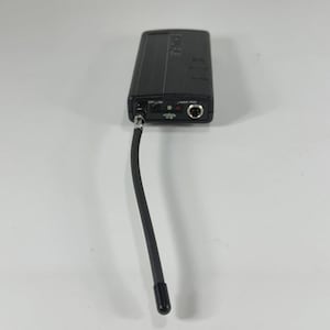 Puede incluir: Un transmisor de micr&oacute;fono inal&aacute;mbrico Shure negro con una antena flexible. El dispositivo rectangular tiene un bot&oacute;n de encendido, una entrada de audio y una peque&ntilde;a pantalla. La antena se extiende hacia abajo, terminando en una punta negra.