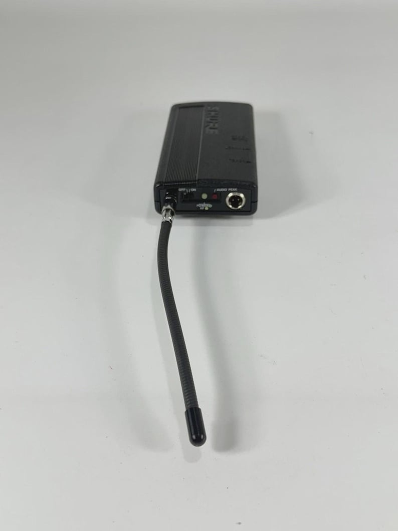 Puede incluir: Un transmisor de micr&oacute;fono inal&aacute;mbrico Shure negro con una antena flexible. El dispositivo rectangular tiene un bot&oacute;n de encendido, una entrada de audio y una peque&ntilde;a pantalla. La antena se extiende hacia abajo, terminando en una punta negra.