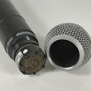 以下が含まれることがあります： 銀色のメッシュグリルが取り外された黒いShure SM58マイク。マイク本体にはブランド名とモデル番号が白で印刷されています。内部のカプセルが見えており、質感のある茶色のフォームフィルターと小さな白いラベルが確認できます。