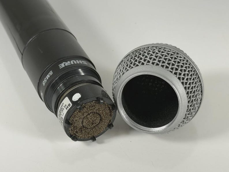 以下が含まれることがあります： 銀色のメッシュグリルが取り外された黒いShure SM58マイク。マイク本体にはブランド名とモデル番号が白で印刷されています。内部のカプセルが見えており、質感のある茶色のフォームフィルターと小さな白いラベルが確認できます。