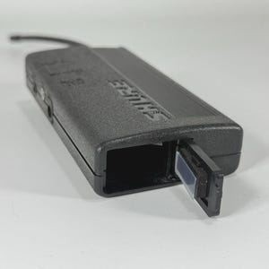 Puede incluir: Un receptor inal&aacute;mbrico Shure negro con un compartimento abierto y una peque&ntilde;a antena. El dispositivo es rectangular con el nombre de la marca en relieve en el lateral. El receptor est&aacute; sobre una superficie blanca.