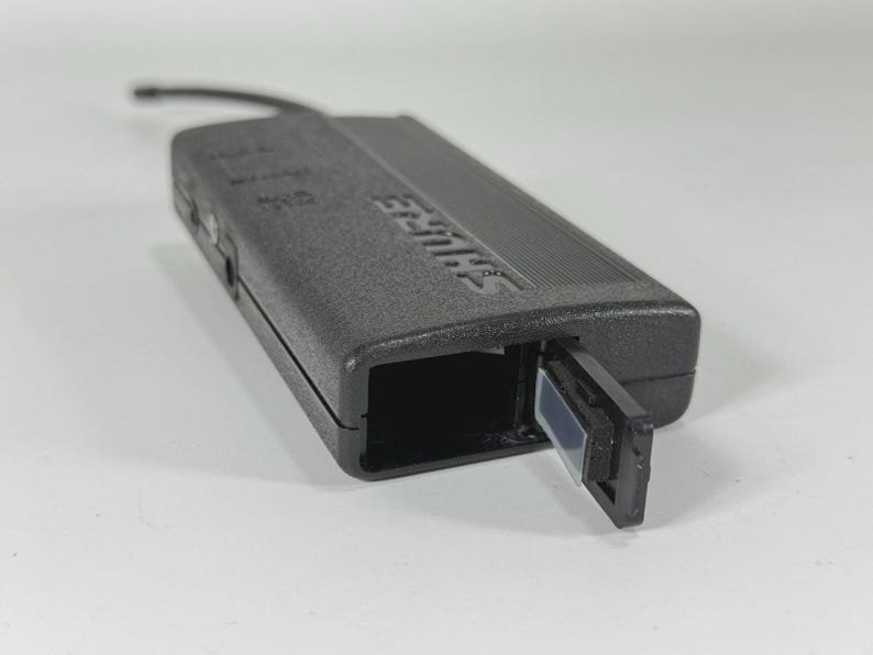 Puede incluir: Un receptor inal&aacute;mbrico Shure negro con un compartimento abierto y una peque&ntilde;a antena. El dispositivo es rectangular con el nombre de la marca en relieve en el lateral. El receptor est&aacute; sobre una superficie blanca.
